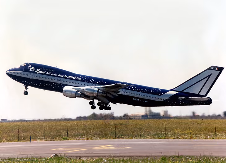 Blue and white Alitalia Boeing 747 airplane taking off from a runway with clear sky.