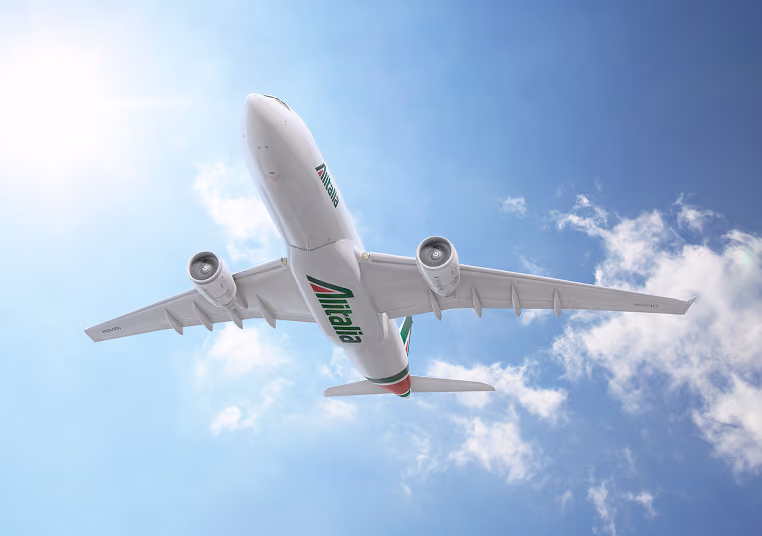 Alitalia airplane flying overhead against a blue sky with sunlight and clouds.